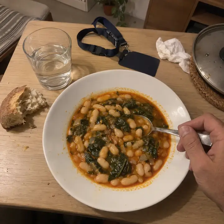 Plato hondo de judías blancas con acelgas con una cuchara y un trozo de pan, en la mesa del comedor por la noche